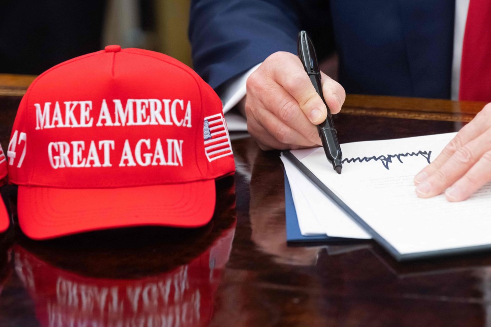 O presidente dos EUA, Donald Trump, assina ordem executiva ao lado de boné com seu slogan ‘Make America Great Again’ — Foto: Saul Loeb/AFP