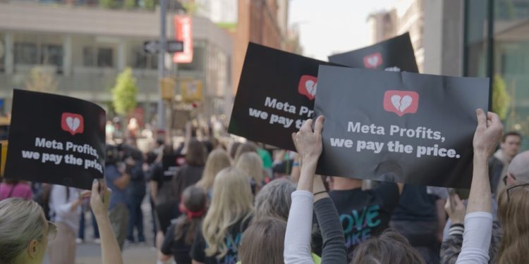 Cena de 'Your attention please', com manifestantes carregando cartazes com os dizeres: 'Meta lucra, nós pagamos o preço' — Foto: Divulgação